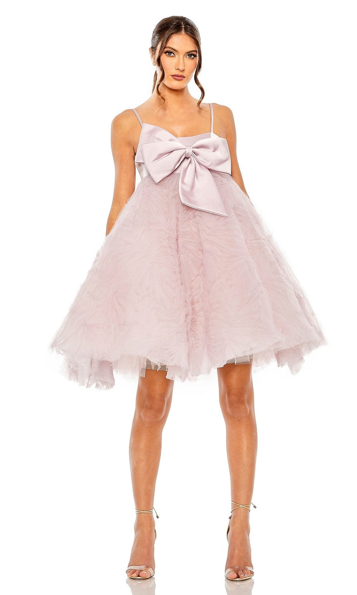 A woman wears the Short Homecoming Dress 20583 by Mac Duggal in rose pink, featuring a large bow on the bodice, sleeveless knee-length design, and silver high-heeled sandals for an elegant yet playful party look.